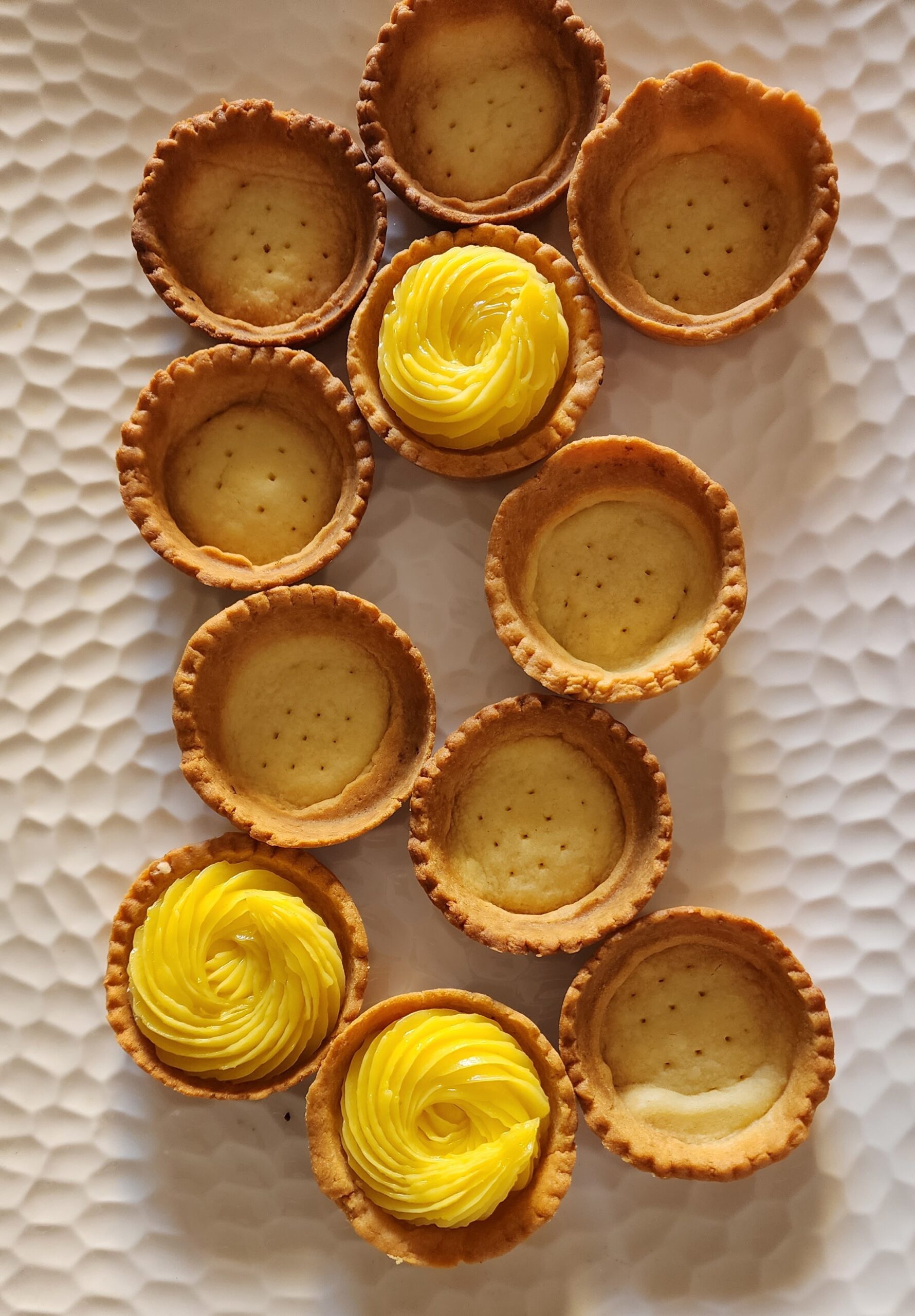 Eleven baked tart shells placed on a white platter with some of the shells filled inside with lemon curd filling 