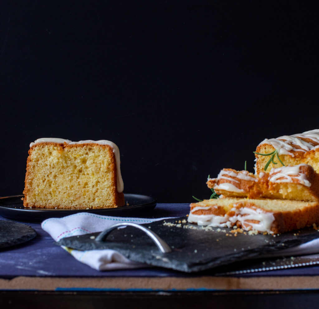 A slice of Lemon Loaf against a black background and sliced cake on right