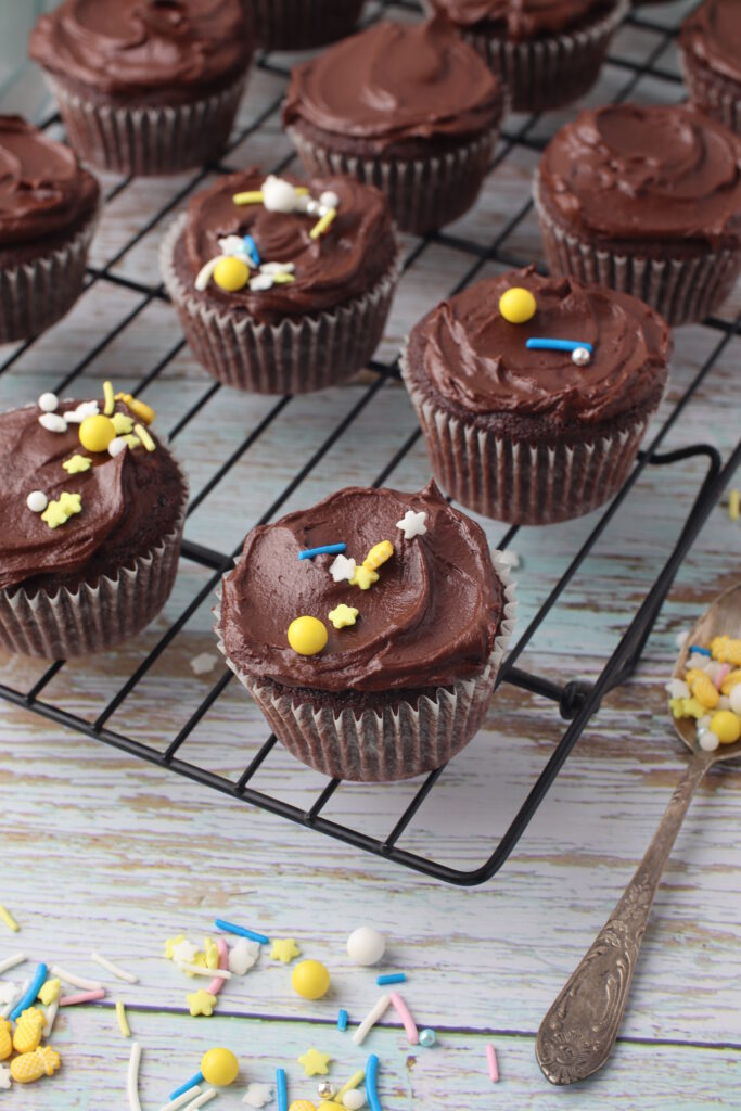Chocolate fudge cupcakes with sprinkles on top placed on a wire rack with a rustic background and scattered sprinkles