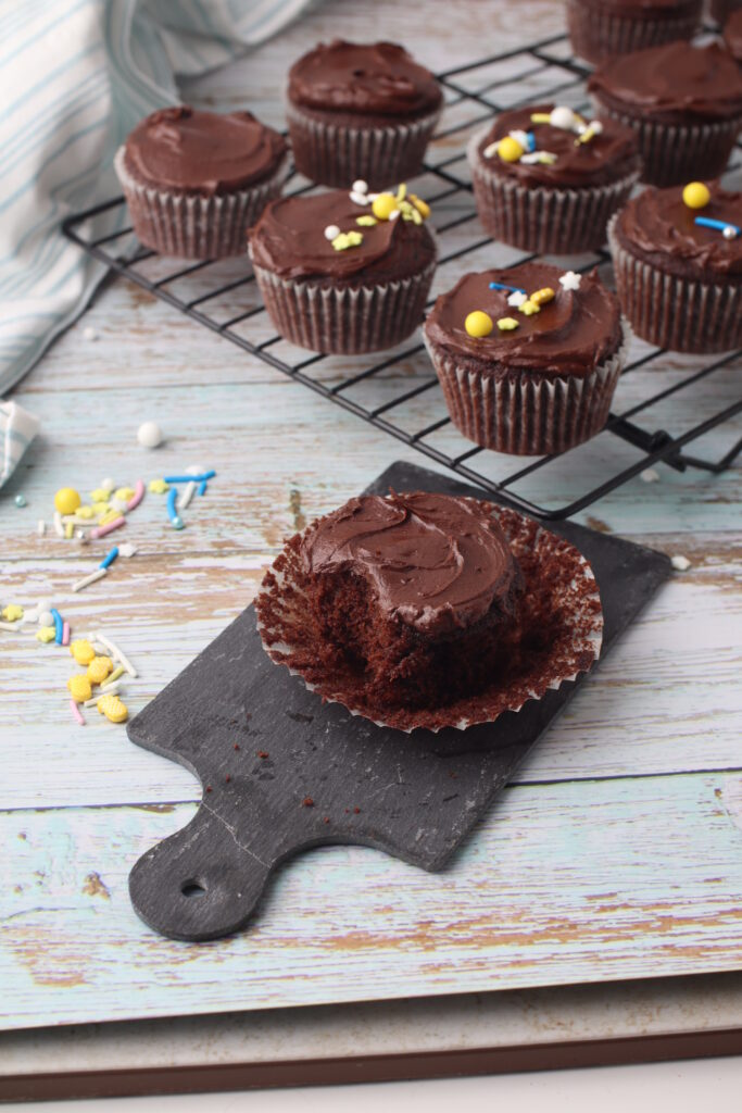 chocolate cupcake, with fudge frosting, one bite taken on a slate with sprinkles around and more frosted cupcakes on the back