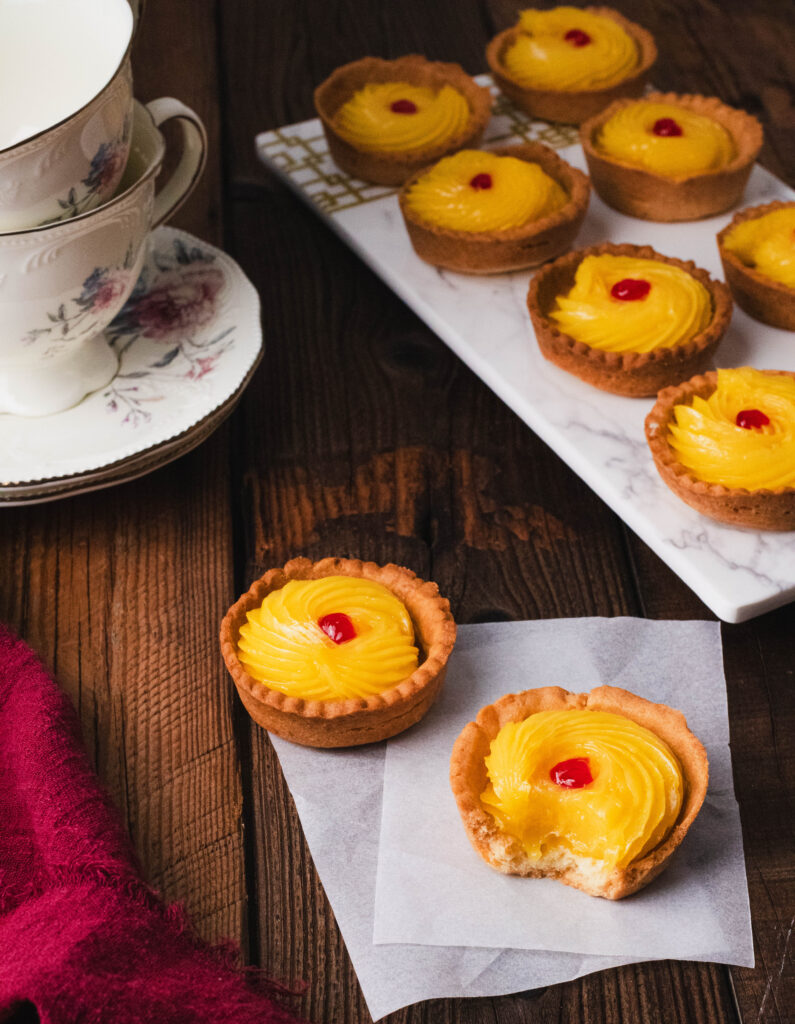 Two lemon tarts (short-crust pastry filled with lemon filling & red jam dot), placed on baking paper, with more tarts in the background along with tea cups on a wooden table.