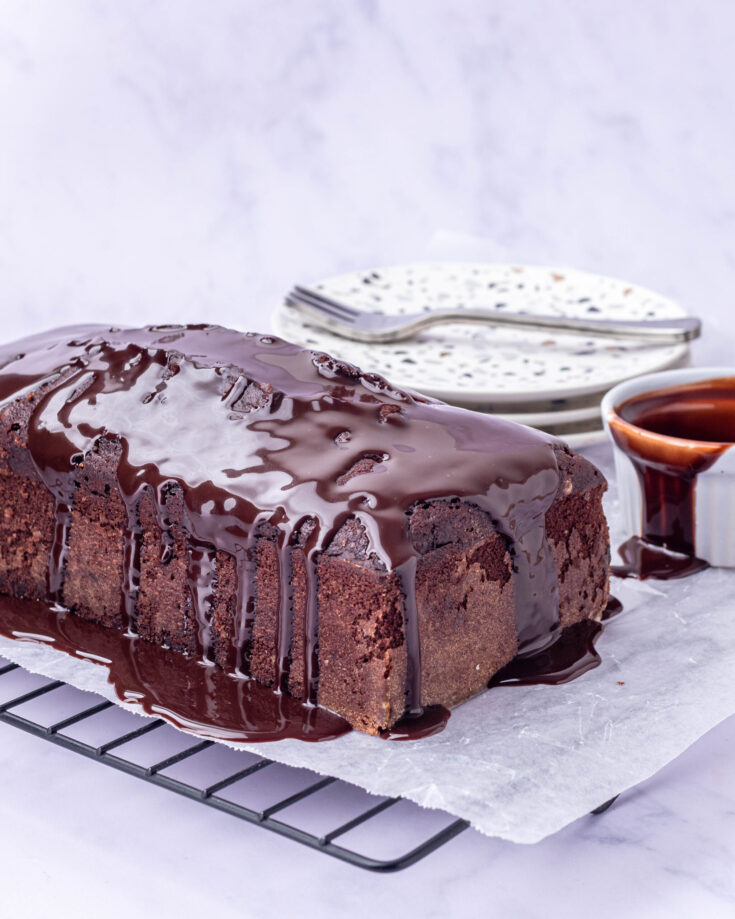 Chocolate Pound Cake (baked in a loaf pan) placed on a wire rack with a few plates & forks, chocolate syrup in a ramekin and a spatula dipped in chocolate syrup in the background