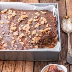Sticky Date Cake in a baking pan, placed on a rustic wooden surface. In the background is butterscotch sauce in a small jar, on the right side is a tablespoon while in front, there is a small dish with soft pitted dates in it.