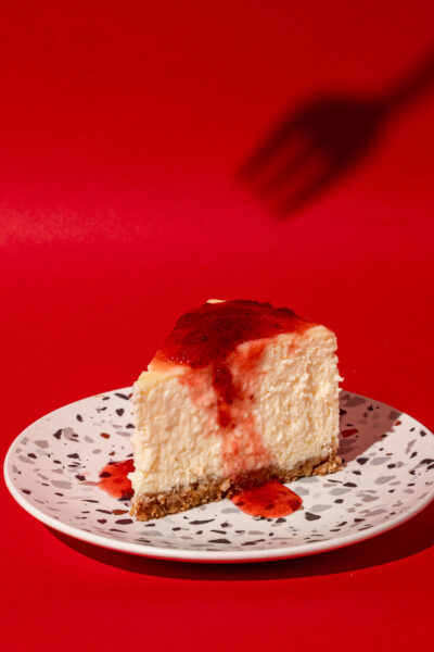 New york cheesecake slice with strawberry compote on a red backdrop and a fork shadow